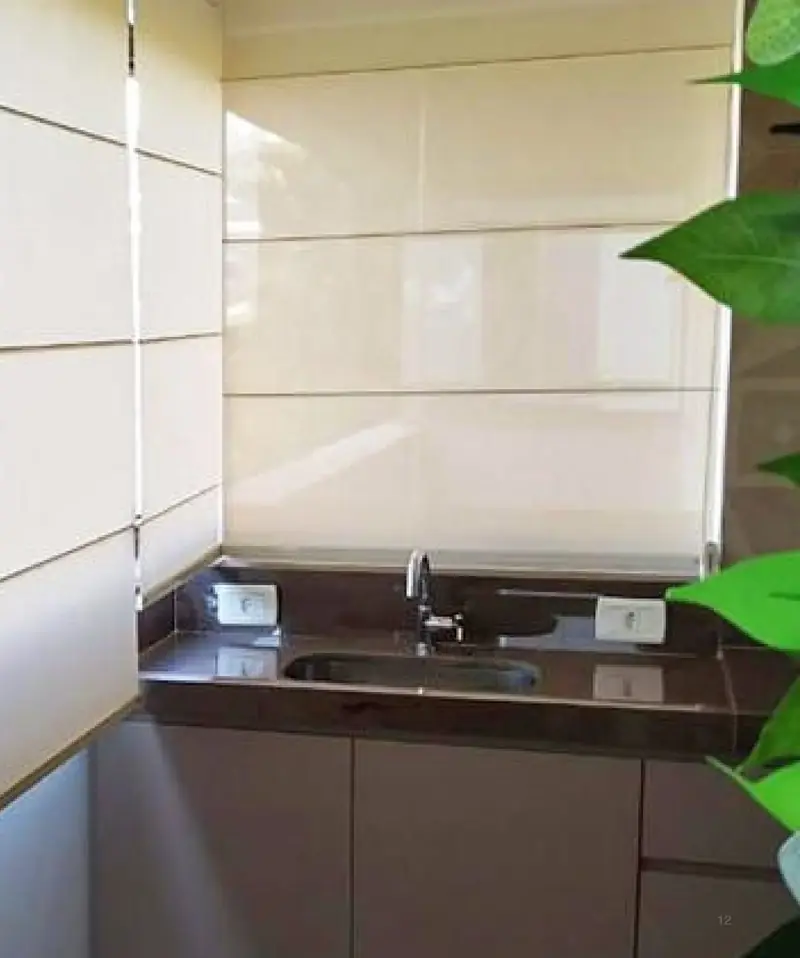 Cortina romana de tecido claro em uma cozinha com bancada escura e plantas ao lado.