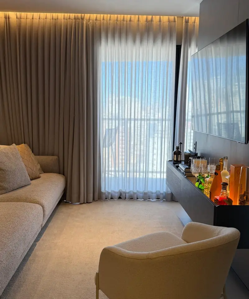Sala com sofá bege e cortina de tecido em tom taupe junto a uma janela com vista para a cidade.