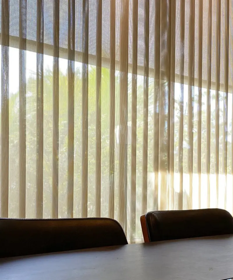 Persiana vertical em tom areia filtrando a luz do jardim em uma sala, com poltronas escuras em primeiro plano.