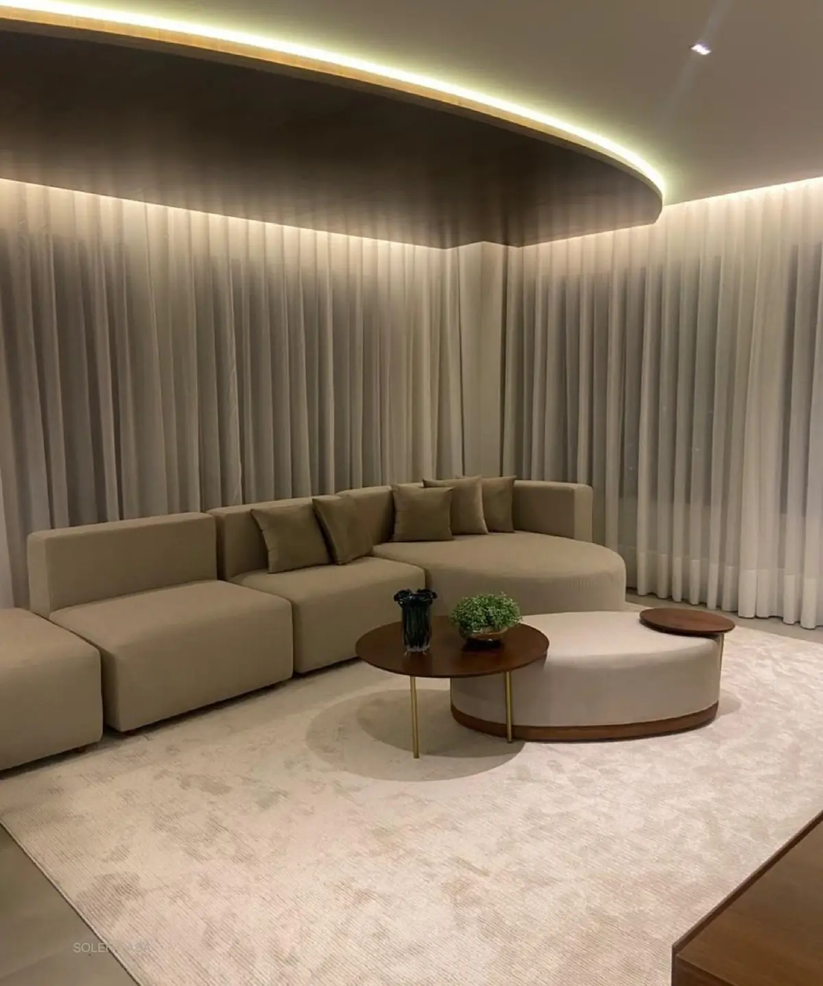Sala de estar com sofá em tons neutros e cortinas de tecido cobrindo as paredes, com iluminação indireta no teto.
